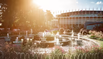 Ein sonnendurchfluteter Garten mit einem zentralen runden Brunnen, umgeben von bunten Blumen und Grünpflanzen. Im Hintergrund ist ein modernes Gebäude mit großen Fenstern und Flaggen auf dem Dach zu sehen. Das Sonnenlicht hüllt die Szene in einen warmen Schein.