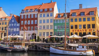 Bunte Gebäude mit roten, gelben, blauen und grünen Fassaden säumen den Nyhavn-Kanal in Kopenhagen. Im Vordergrund liegen mehrere Boote vor Anker, und entlang der Uferpromenade sind unter einem strahlend blauen Himmel Straßencafés zu sehen.