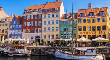 Bunte Gebäude mit roten, gelben, blauen und grünen Fassaden säumen den Nyhavn-Kanal in Kopenhagen. Im Vordergrund liegen mehrere Boote vor Anker, und entlang der Uferpromenade sind unter einem strahlend blauen Himmel Straßencafés zu sehen.