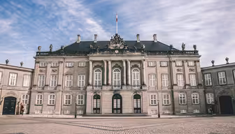 Elegantes, historisches europäisches Gebäude mit symmetrischer Fassade, Bogenfenstern, kunstvollen Skulpturen und einem zentralen Fahnenmast. Im Vordergrund ein gepflasterter Platz unter einem teilweise bewölkten Himmel.