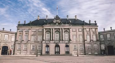 Elegantes, historisches europäisches Gebäude mit symmetrischer Fassade, Bogenfenstern, kunstvollen Skulpturen und einem zentralen Fahnenmast. Im Vordergrund ein gepflasterter Platz unter einem teilweise bewölkten Himmel.