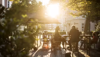 An einem sonnigen Tag sitzen Menschen in einem Straßencafé. Die Sonne wirft einen warmen Schein durch die Bäume. Tische und Stühle stehen auf einer gepflasterten Fläche, und die Atmosphäre ist entspannt und einladend.