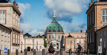 Zwei Personen fahren mit dem Fahrrad auf einem historischen Platz in Kopenhagen, Dänemark. Die Szene wird von klassischen Gebäuden und einer Kirche mit einer großen grünen Kuppel unter einem teilweise bewölkten Himmel eingerahmt. Im Vordergrund sind Kopfsteinpflaster und Poller zu sehen.