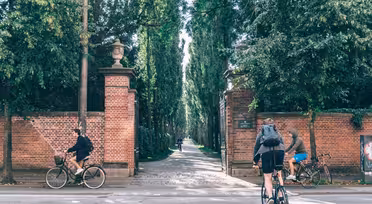 Zwei Radfahrer fahren an roten Backsteinsäulen vorbei, die den Eingang zu einem von Bäumen gesäumten Weg markieren. Ein Radfahrer fährt auf den Eingang zu, der andere entfernt sich. Dichtes Grün säumt den Weg und schafft eine schattige, ruhige Atmosphäre.