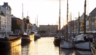 Boote reihen sich entlang eines Kanals in einem sonnenbeschienenen Stadtgebiet auf. Historische Gebäude und ein warmer Sonnenuntergang im Hintergrund prägen das Bild. Ruhiges Wasser spiegelt die Gebäude und Boote und schafft so eine heitere Atmosphäre.