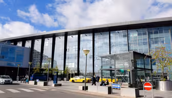 Ein modernes Flughafenterminal mit Glasfassade. Draußen parken mehrere gelbe Taxis und einige Passanten. Im Hintergrund ist ein blauer Himmel mit vereinzelten Wolken zu sehen. Im Vordergrund sind ein Zebrastreifen und Verkehrsschilder zu sehen.