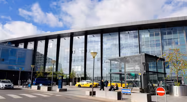 Ein modernes Flughafenterminal mit Glasfassade. Draußen parken mehrere gelbe Taxis und einige Passanten. Im Hintergrund ist ein blauer Himmel mit vereinzelten Wolken zu sehen. Im Vordergrund sind ein Zebrastreifen und Verkehrsschilder zu sehen.