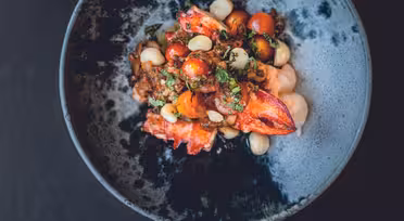 Ein Gourmetgericht auf einem blauen Teller mit gemischten Kirschtomaten, weißen Bohnen, Kräutern und Hummerfleisch, garniert mit frischem Grün. Die Präsentation ist kunstvoll und farbenfroh, vor einem dunklen Hintergrund.