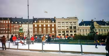 Menschen laufen auf einer Eisbahn Schlittschuh, umgeben von historischen Gebäuden im europäischen Stil, unter einem bewölkten Himmel. Rechts ist eine Statue zu sehen, und mehrere Zuschauer beobachten die Schlittschuhläufer.