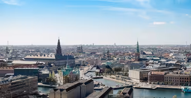 Luftaufnahme einer Stadtlandschaft mit historischen Gebäuden, darunter Türme und Dächer unter einem klaren blauen Himmel. Ein Fluss mit Brücken fließt durch die Szene, umgeben von einer Mischung aus alter und moderner Architektur.