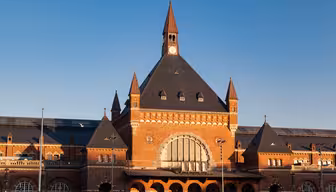 Ein historischer Bahnhof mit großer Backsteinfassade, einem zentralen Bogenfenster und einem markanten Uhrenturm. Das Gebäude ist mit symmetrischen Spitzdächern vor einem klaren blauen Himmel geschmückt.