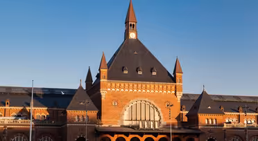 Ein historischer Bahnhof mit großer Backsteinfassade, einem zentralen Bogenfenster und einem markanten Uhrenturm. Das Gebäude ist mit symmetrischen Spitzdächern vor einem klaren blauen Himmel geschmückt.