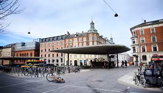 Ein Platz mit einer einzigartigen kreisförmigen Dachkonstruktion in der Mitte, umgeben von Fahrradständern. Im Hintergrund sind historische Gebäude mit roten und beigen Backsteinfassaden zu sehen. Der Himmel ist bewölkt, und einige Menschen gehen in der Nähe spazieren.