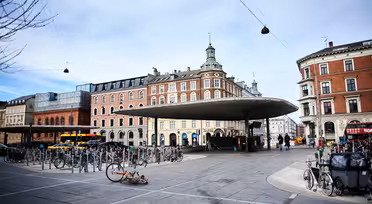 Ein Platz mit einer einzigartigen kreisförmigen Dachkonstruktion in der Mitte, umgeben von Fahrradständern. Im Hintergrund sind historische Gebäude mit roten und beigen Backsteinfassaden zu sehen. Der Himmel ist bewölkt, und einige Menschen gehen in der Nähe spazieren.