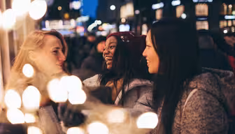 Drei Frauen lächeln und unterhalten sich in einer lebhaften nächtlichen Umgebung, geschmückt mit festlichen Lichtern. Sie sind warm gekleidet, was auf die kalte Jahreszeit hindeutet. Der unscharfe Hintergrund zeigt helle, unscharfe Lichter und eine geschäftige Menschenmenge.