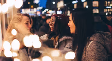 Drei Frauen lächeln und unterhalten sich in einer lebhaften nächtlichen Umgebung, geschmückt mit festlichen Lichtern. Sie sind warm gekleidet, was auf die kalte Jahreszeit hindeutet. Der unscharfe Hintergrund zeigt helle, unscharfe Lichter und eine geschäftige Menschenmenge.