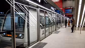 Eine U-Bahn hält in einer modernen U-Bahn-Station. Menschen stehen an den Glasbarrieren, manche steigen ein, andere warten. Auffällige Deckenleuchten erhellen den Bahnsteig, digitale Anzeigen zeigen Zuginformationen an.