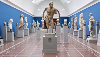 Eine Museumsgalerie präsentiert verschiedene antike Steinskulpturen auf Sockeln. Der Raum hat eine blaue Wand und eine hohe gewölbte Decke mit Oberlichtern, die eine helle und luftige Atmosphäre schaffen. Besucher betrachten die Skulpturen.