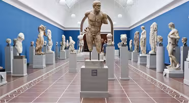 Eine Museumsgalerie präsentiert verschiedene antike Steinskulpturen auf Sockeln. Der Raum hat eine blaue Wand und eine hohe gewölbte Decke mit Oberlichtern, die eine helle und luftige Atmosphäre schaffen. Besucher betrachten die Skulpturen.