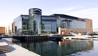 Modernes Einkaufszentrum am Wasser mit großen Glasfenstern und einem zylindrischen Eingang mit der Aufschrift „Fisketorvet“. Umgeben von stillem Wasser, Holzstegen und klarem Himmel ist die Spiegelung des Gebäudes im Wasser sichtbar.