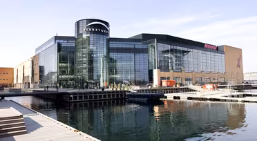 Modernes Einkaufszentrum am Wasser mit großen Glasfenstern und einem zylindrischen Eingang mit der Aufschrift „Fisketorvet“. Umgeben von stillem Wasser, Holzstegen und klarem Himmel ist die Spiegelung des Gebäudes im Wasser sichtbar.