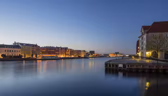 Eine ruhige Uferszene in der Abenddämmerung: Der ruhige Fluss spiegelt die Lichter der Stadt. Historische Gebäude säumen das Ufer, darüber ein klarer, tiefblauer Himmel. Die Straßenlaternen verleihen der Stadtlandschaft einen warmen Glanz.