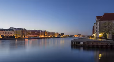 Eine ruhige Uferszene in der Abenddämmerung: Der ruhige Fluss spiegelt die Lichter der Stadt. Historische Gebäude säumen das Ufer, darüber ein klarer, tiefblauer Himmel. Die Straßenlaternen verleihen der Stadtlandschaft einen warmen Glanz.