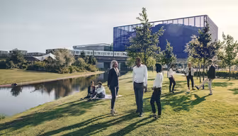 Menschen haben sich auf einer Rasenfläche mit Bäumen an einem Teich versammelt. Im Hintergrund sind ein modernes Glasgebäude und Bahngleise zu sehen. Einige stehen in Businesskleidung, andere sitzen und genießen den sonnigen Tag.