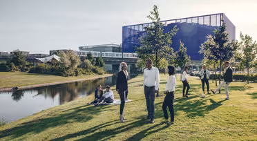 Menschen haben sich auf einer Rasenfläche mit Bäumen an einem Teich versammelt. Im Hintergrund sind ein modernes Glasgebäude und Bahngleise zu sehen. Einige stehen in Businesskleidung, andere sitzen und genießen den sonnigen Tag.