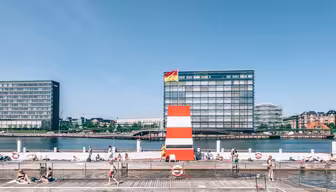 Menschen genießen einen sonnigen Tag in einem Freibad mit einem großen rot-weißen Sprungturm. Im Hintergrund sind moderne Gebäude zu sehen, und Menschen liegen am Wasser. Der Himmel ist klar und blau.