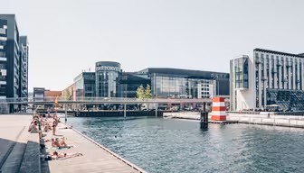 Menschen sonnen sich auf einer Holzplattform am Kanal. Moderne Gebäude, darunter ein Einkaufszentrum mit Glasfassade, säumen das Wasser. Eine Brücke überspannt den Kanal, und im Wasser ist eine gestreifte Boje zu sehen. Der Himmel ist klar und sonnig.