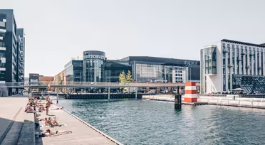 Menschen sonnen sich auf einer Holzplattform am Kanal. Moderne Gebäude, darunter ein Einkaufszentrum mit Glasfassade, säumen das Wasser. Eine Brücke überspannt den Kanal, und im Wasser ist eine gestreifte Boje zu sehen. Der Himmel ist klar und sonnig.