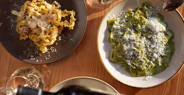 Zwei Teller Pasta auf einem Holztisch: einer mit gewellten Nudeln und Kräutern, der andere mit grünen Spinatnudeln, beide mit geriebenem Käse garniert. Aus einer Flasche fließt Flüssigkeit in ein Glas daneben.