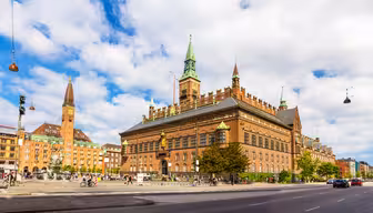 Eine lebendige Szene des Kopenhagener Rathauses mit seiner roten Backsteinarchitektur und den grünen Türmen unter einem leicht bewölkten Himmel. Im Vordergrund spazieren Menschen in der Nähe eines Brunnens, und die belebte Straße verleiht der historischen Kulisse Leben.
