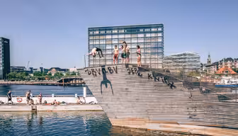 Menschen stehen auf einem hölzernen Sprungturm am Ufer, im Hintergrund eine Stadtlandschaft mit modernen Gebäuden. Der klare Himmel und das sonnige Wetter deuten auf einen warmen Tag hin. An der Seite des Sprungturms sind Graffiti zu sehen.