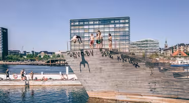 Menschen stehen auf einem hölzernen Sprungturm am Ufer, im Hintergrund eine Stadtlandschaft mit modernen Gebäuden. Der klare Himmel und das sonnige Wetter deuten auf einen warmen Tag hin. An der Seite des Sprungturms sind Graffiti zu sehen.