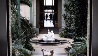 Blick durch ein Bogenfenster auf einen Innengarten mit einer zentralen Statue einer liegenden Figur. Tropische Pflanzen säumen den Weg, der zu einer entfernten Tür führt, in der die Silhouetten zweier Personen stehen.