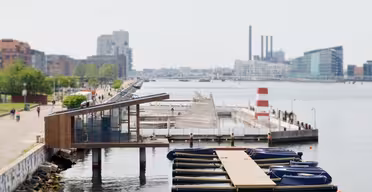 Eine moderne Uferpromenade mit einem kleinen Yachthafen und mehreren angedockten Booten. Ein Holzgebäude ragt über das Wasser, und in der Ferne ist ein rot-weißer Turm zu sehen. An einem klaren Tag flanieren Menschen entlang der baumgesäumten Promenade.