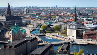 Luftaufnahme einer Stadtlandschaft mit Kanälen, historischen Gebäuden mit Türmen und moderner Architektur. Die Szene umfasst Brücken über dem Wasser und grüne Baumflächen unter einem klaren blauen Himmel.