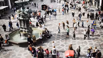 Ein belebter Stadtplatz mit Menschen, die um einen Brunnen herum spazieren und sich versammeln. Der gemusterte Bürgersteig des Platzes ist von historischen Gebäuden umgeben. Man sieht Menschen flanieren, fotografieren und sitzen. Helles und geschäftiges Stadtbild.