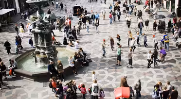 Ein belebter Stadtplatz mit Menschen, die um einen Brunnen herum spazieren und sich versammeln. Der gemusterte Bürgersteig des Platzes ist von historischen Gebäuden umgeben. Man sieht Menschen flanieren, fotografieren und sitzen. Helles und geschäftiges Stadtbild.