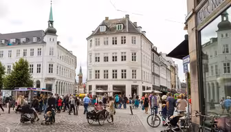 Ein belebter europäischer Stadtplatz mit Menschen, die spazieren gehen, Rad fahren oder in Straßencafés sitzen. Historische Gebäude mit klassischer Architektur umgeben den Platz. Im Hintergrund ist ein Kirchturm zu sehen, im Vordergrund eine Kopfsteinpflasterstraße.