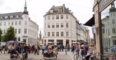Ein belebter europäischer Stadtplatz mit Menschen, die spazieren gehen, Rad fahren oder in Straßencafés sitzen. Historische Gebäude mit klassischer Architektur umgeben den Platz. Im Hintergrund ist ein Kirchturm zu sehen, im Vordergrund eine Kopfsteinpflasterstraße.