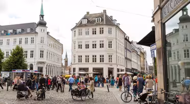 Ein belebter europäischer Stadtplatz mit Menschen, die spazieren gehen, Rad fahren oder in Straßencafés sitzen. Historische Gebäude mit klassischer Architektur umgeben den Platz. Im Hintergrund ist ein Kirchturm zu sehen, im Vordergrund eine Kopfsteinpflasterstraße.