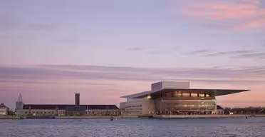 Ein modernes Opernhaus mit flachem, überhängendem Dach liegt bei Sonnenuntergang am Wasser. Der Himmel ist in rosa und violette Töne getaucht. Daneben steht ein langes, niedriges historisches Gebäude mit einem Turm.
