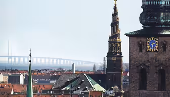 Stadtlandschaft mit einem kunstvoll gemusterten Kirchturm und einer Glocke, Gebäuden mit roten Dächern und einer entfernten Brücke, die sich unter einem klaren Himmel über das Wasser spannt.