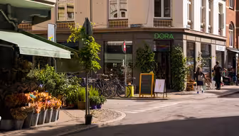 Eine sonnige Straßenecke. Links ein Blumenladen mit Pflanzen und Sträußen. Fahrräder parken in der Nähe eines Ladens namens „DORA“. Menschen gehen in der Nähe spazieren, und auf einer Kreidetafel ist eine Eistüte abgebildet. Die Gebäude sind mit Grünpflanzen geschmückt.