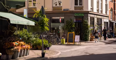 Eine sonnige Straßenecke. Links ein Blumenladen mit Pflanzen und Sträußen. Fahrräder parken in der Nähe eines Ladens namens „DORA“. Menschen gehen in der Nähe spazieren, und auf einer Kreidetafel ist eine Eistüte abgebildet. Die Gebäude sind mit Grünpflanzen geschmückt.