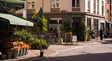 Eine sonnige Straßenecke. Links ein Blumenladen mit Pflanzen und Sträußen. Fahrräder parken in der Nähe eines Ladens namens „DORA“. Menschen gehen in der Nähe spazieren, und auf einer Kreidetafel ist eine Eistüte abgebildet. Die Gebäude sind mit Grünpflanzen geschmückt.