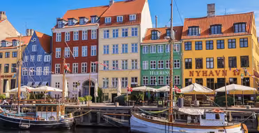 Bunte Gebäude säumen die Uferpromenade im Kopenhagener Nyhavn mit ihren roten, gelben und grünen Fassaden. Boote liegen am Dock, und am Wasser stehen Straßencafés mit Sonnenschirmen. Der Himmel ist klar und blau.